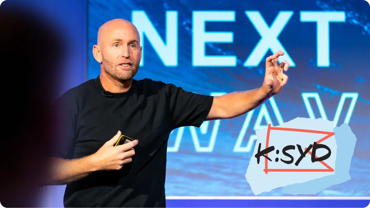 A person speaking on stage with "NEXT WAVE" text in the background and a "K:SYD" logo overlay.