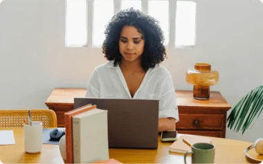 Woman working on a laptop at a wooden desk with books, a mug, and a plant nearby.