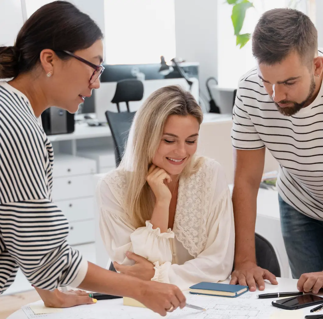 Three people collaborating around a table, looking at documents and discussing ideas in an office setting.