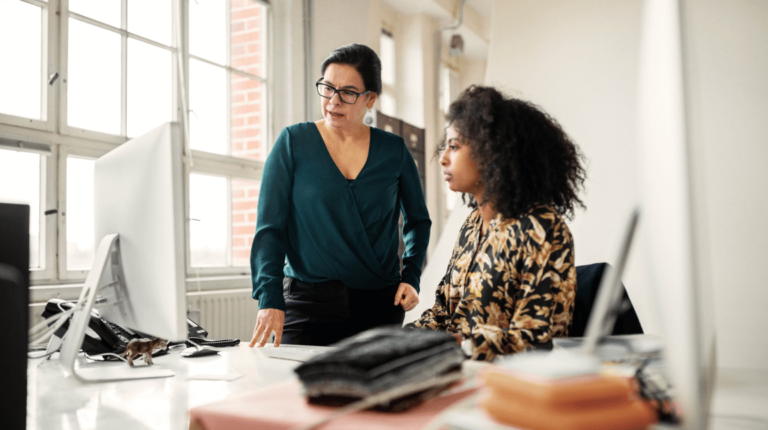 a woman sitting next to a woman standing, both looking at a monitor