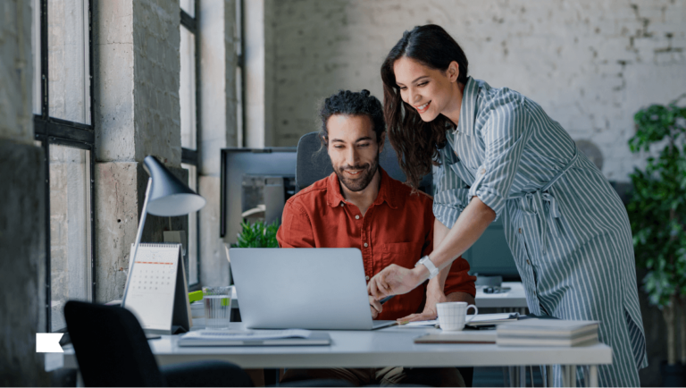 Person sitting at desk on laptop with another person standing next to them pointing at screen