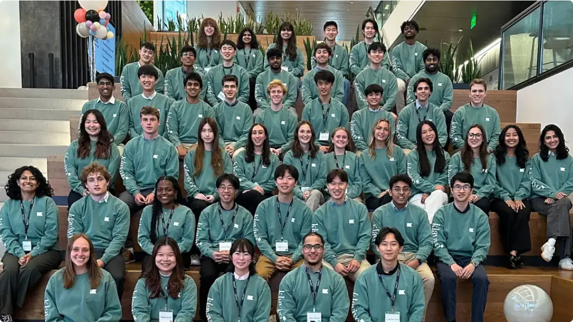 A group of interns wearing matching green sweatshirts, seated on steps in a modern building, posing for a group photo.