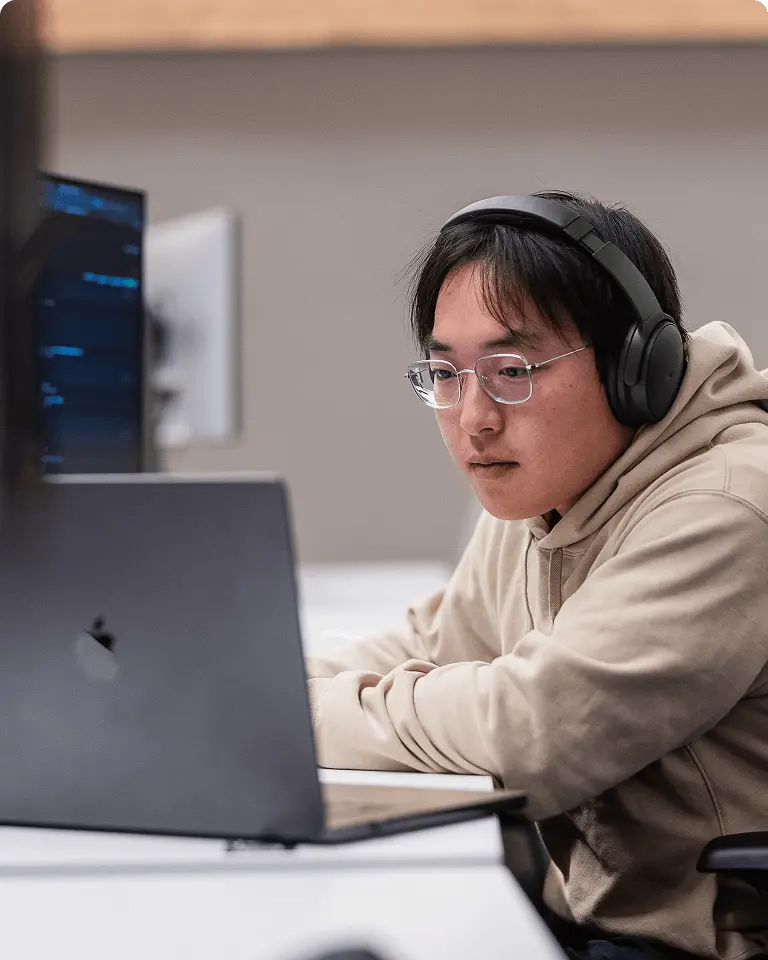 Person wearing headphones and a hoodie, focused on a laptop in an office setting.